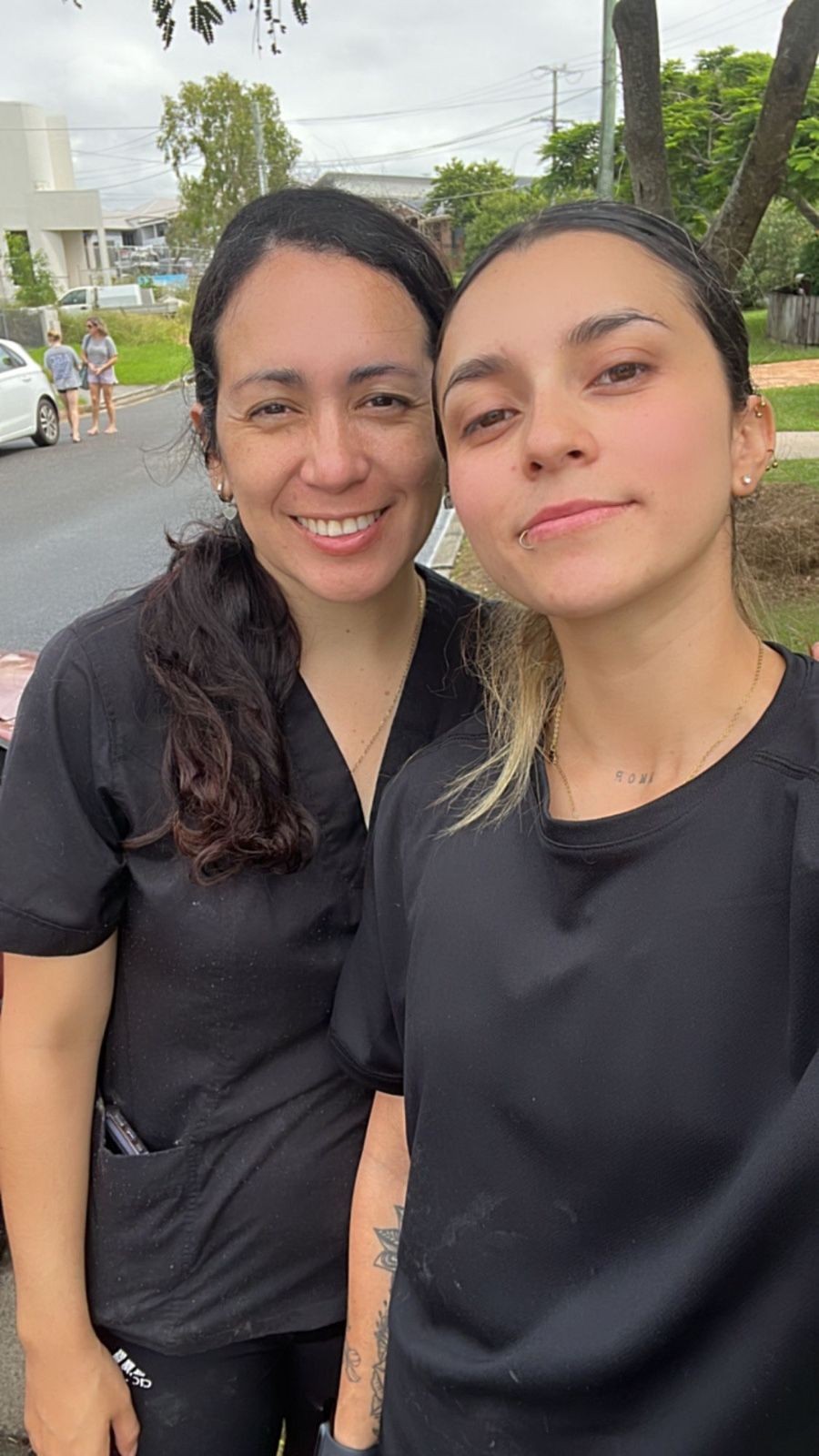 Two people in black shirts taking a selfie outdoors, with trees and cars in the background.